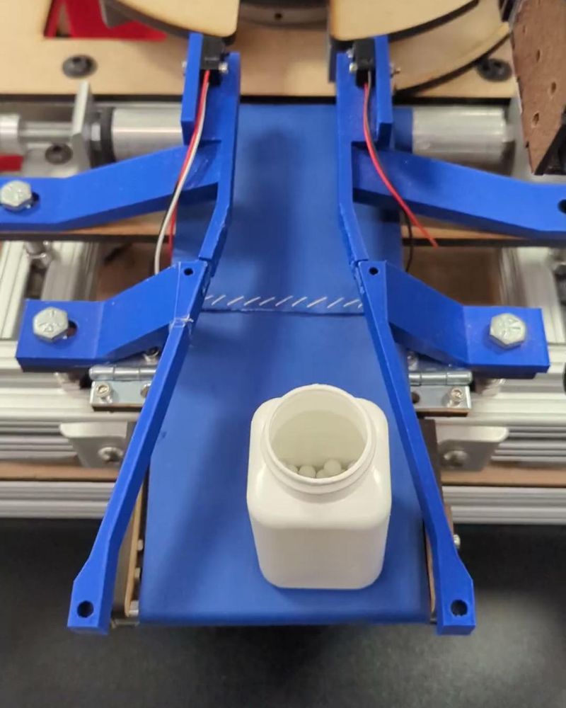 Top-down view of the blue 3D-printed conveyor and guide rails carrying a pill bottle toward the rotary table
